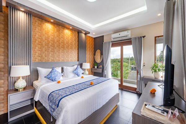 Spacious Balinese-style bedroom with king bed adorned in white linens and blue accents, wooden paneling, tropical garden view through sliding doors, and modern TV.